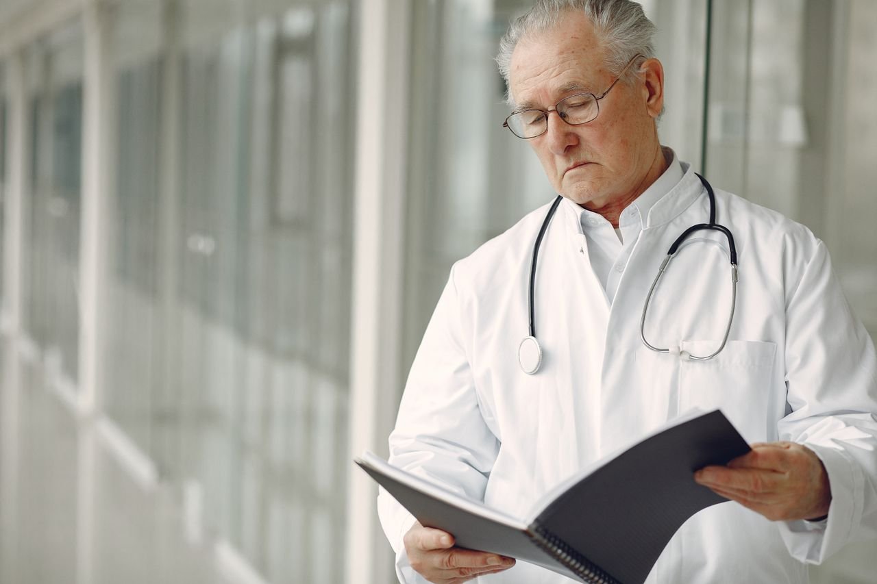 Crop concentrated professional senior male physician in white robe with stethoscope standing in modern hospital hallway and reading medical case records in daylight