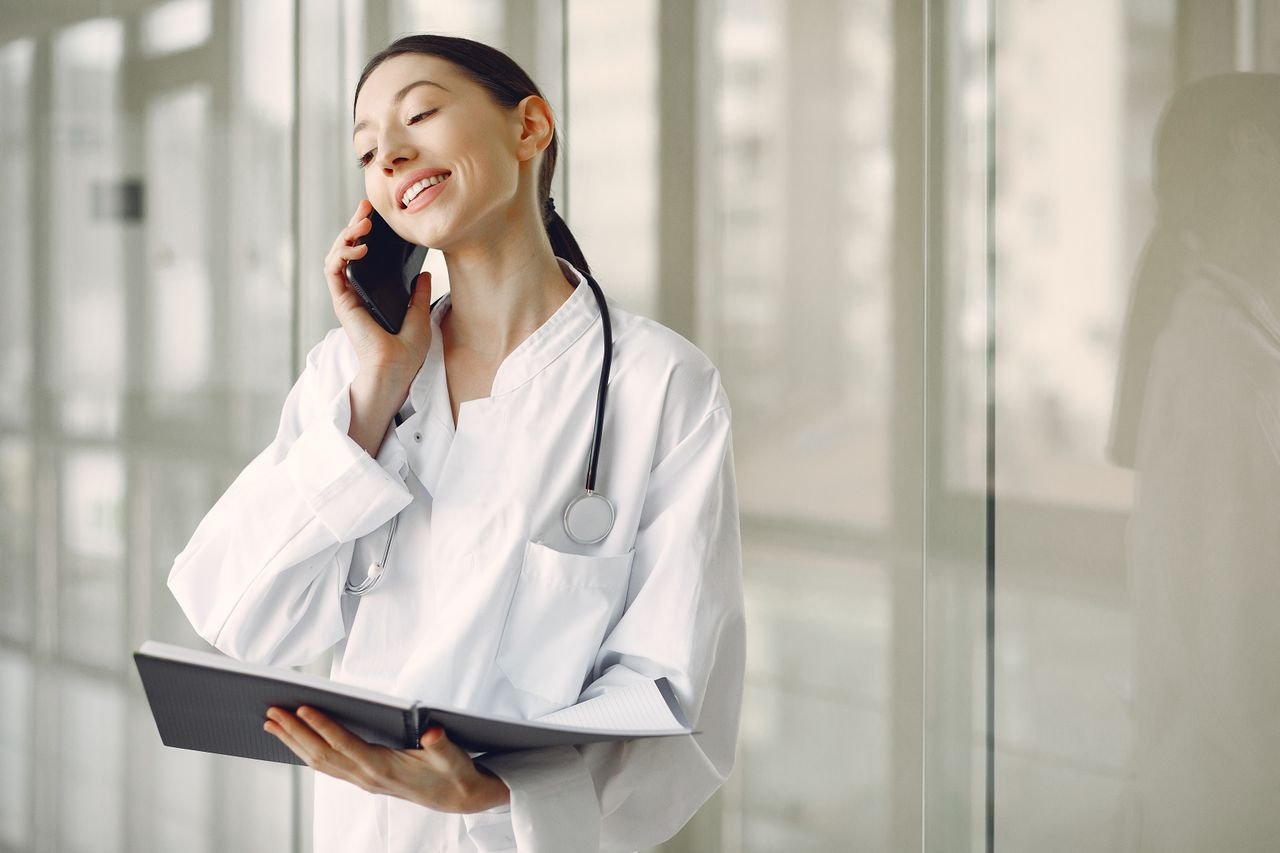 The Art of Drawing Readers In: Your attractive post title goes here Smiling ethnic female doctor in medical uniform with stethoscope and notebook talking on smartphone while standing in glass hospital corridor